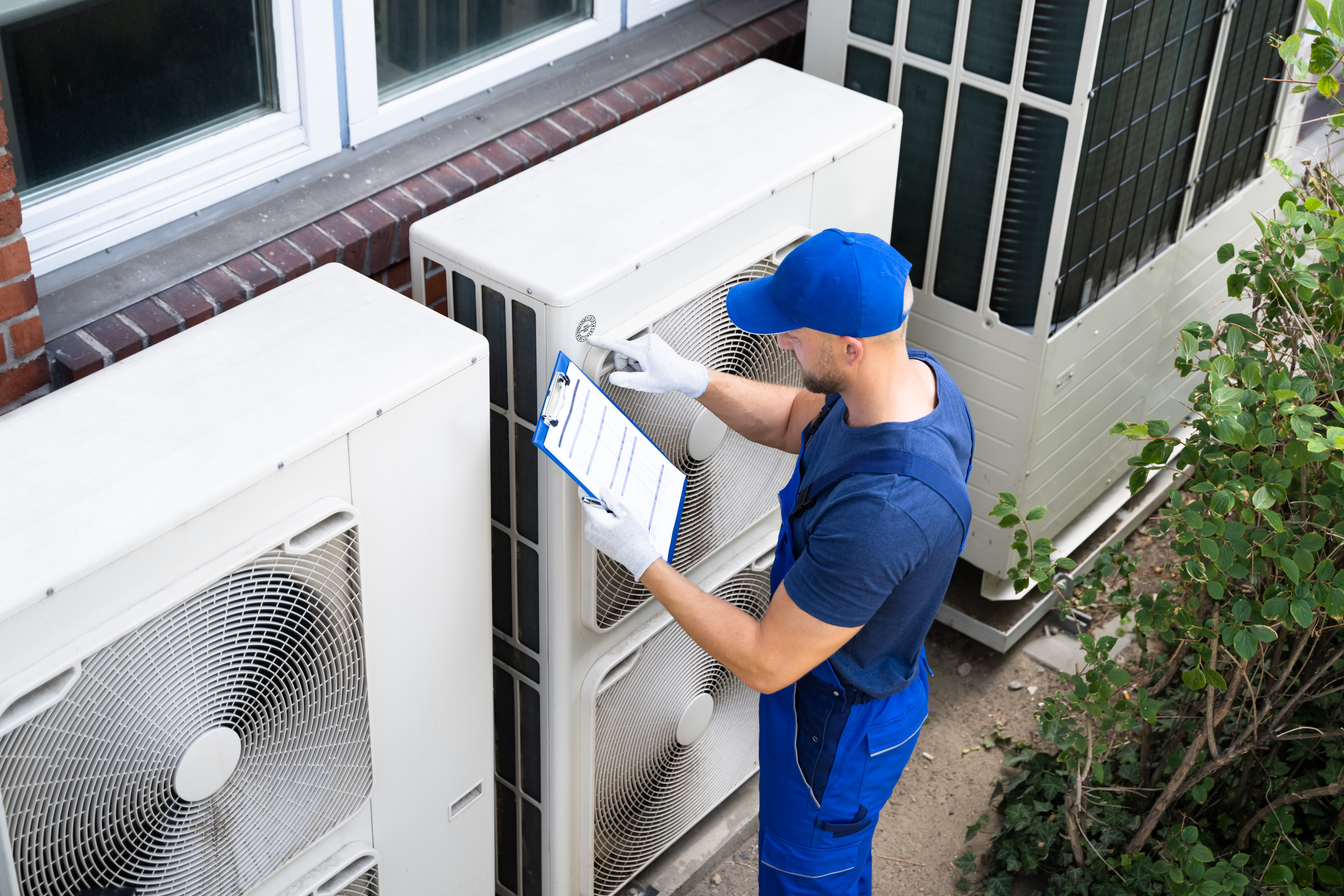 A male worker in a blue uniform uses a checklist and clipboard to perform preventive maintenance on commercial HVAC equipment outdoors. 