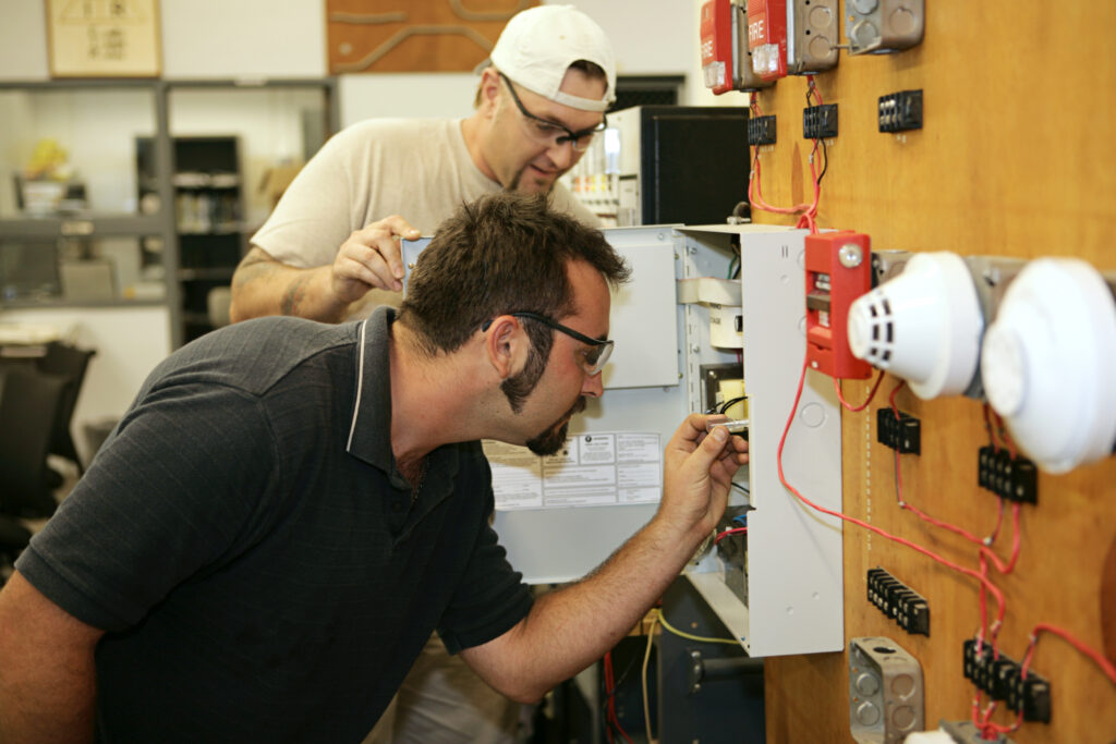 Two workers use tools to check a fuse box while performing school maintenance. 
