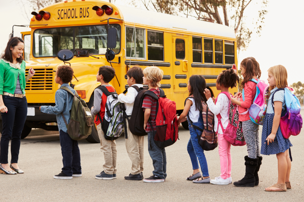 Elementary school students line up in front of their teacher before filing into a school bus.